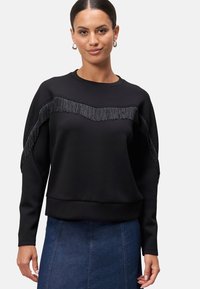 Černá mikina se třásněmi na hrudi, kulatým výstřihem a dlouhými rukávy. Hladká textura, volný střih.