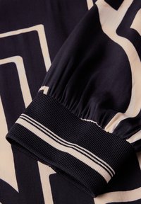 Blusa in tessuto navy con maniche a elastico, caratterizzata da un motivo geometrico crema e un accento a righe sul polsino. Texture liscia.