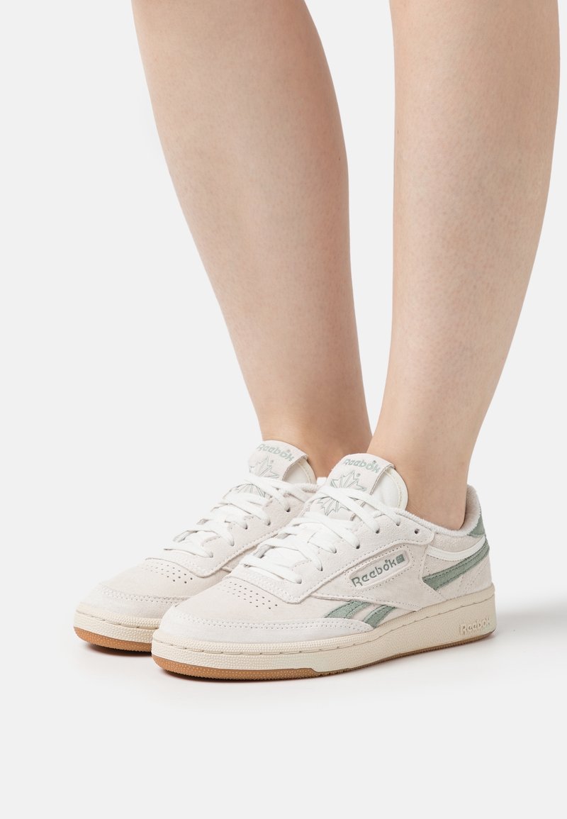 Jambes portant des baskets blanches et gris clair Reebok avec des semelles en gomme, debout sur un fond blanc uni.