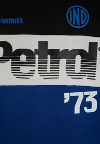 T-shirt blu e nera con il logo "Petrol" in bold nero, testo bianco "'73" e un emblema circolare blu. Il tessuto appare liscio e simile al cotone.