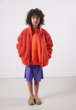 Bambino con capelli ricci che indossa una giacca in pile arancione oversize, una felpa arancione, pantaloncini viola e scarpe slip-on color sabbia, in piedi contro un muro bianco.