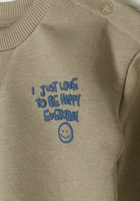 Bézs pulóver kék hímzett szöveggel: "Egyszerűen szeretem minden nap boldognak lenni" mosolygós arccal. Sima textúra, bordázott nyakkivágás és vállgomb.