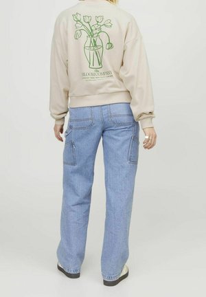 Person trägt einen beigen Pullover mit einem grünen Blumenvase-Grafik und Text, kombiniert mit hellblauen, locker sitzenden Jeans und weißen Schuhen.