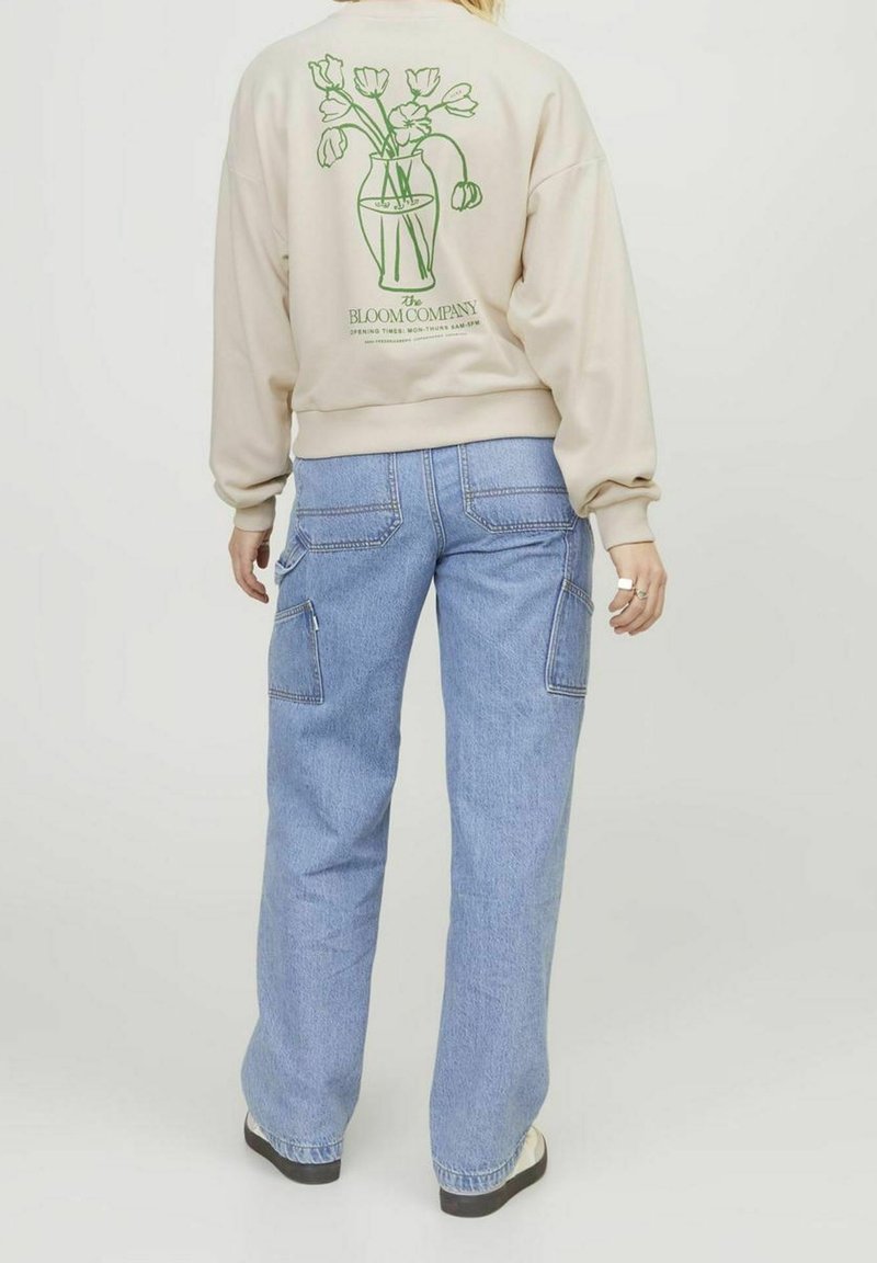 Person wearing beige sweatshirt with green floral vase graphic and text, paired with light blue loose-fitting jeans and white shoes.