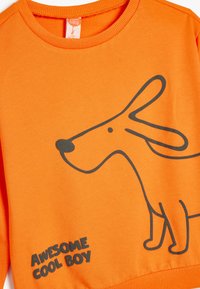 Pomarańczowa bluza z czarną grafiką psa i napisem "SUPER FAJNY CHŁOPAK." Posiada okrągły ściągacz oraz miękką, gładką teksturę materiału.