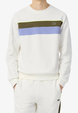 Homme portant un sweat-shirt blanc avec des bandes olive et bleues sur la poitrine et un pantalon blanc assorti, les mains dans les poches.