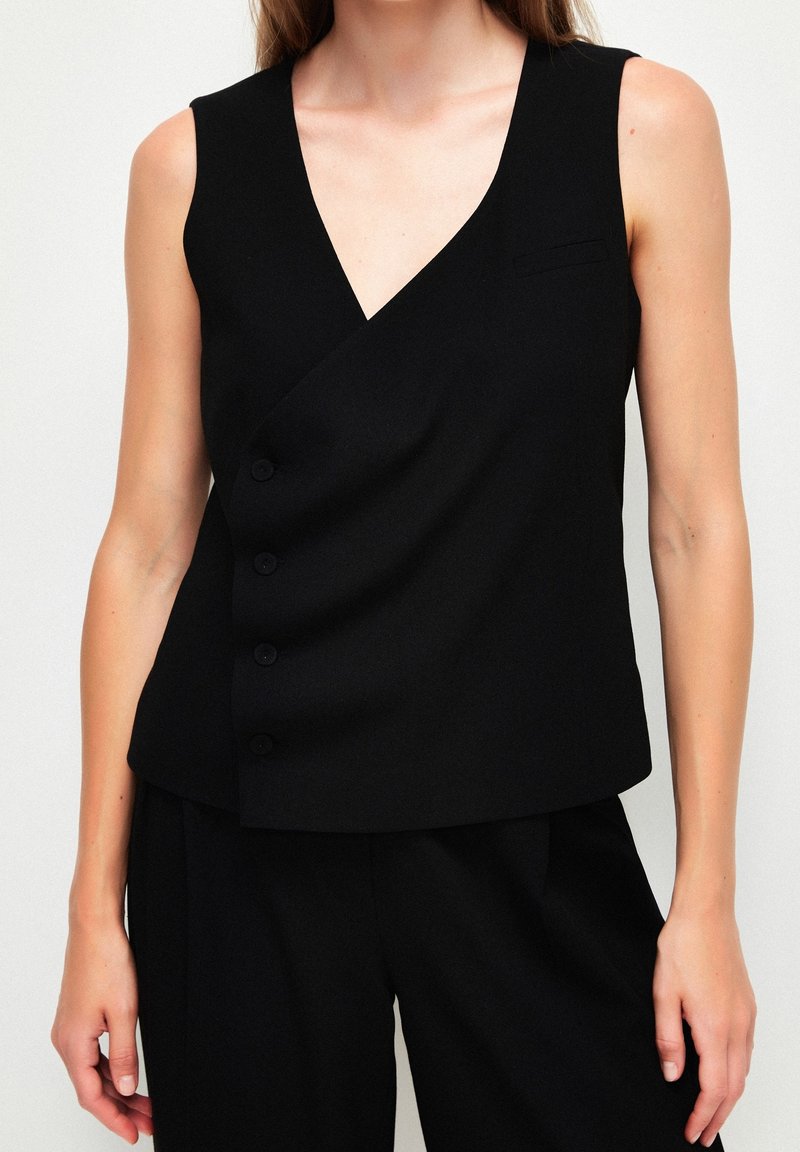 Femme portant un gilet noir sans manches à col en V boutonné et un pantalon noir assorti sur un fond uni.