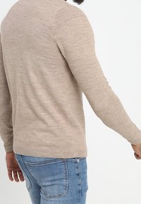 Beżowy dziergany sweter, tkanina o fakturze, dopasowany fason, długie rękawy, zaokrąglony dół, noszony z jasnoniebieskimi dżinsami, styl casualowy.