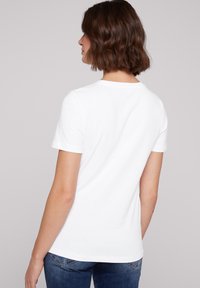 Camiseta blanca de manga corta con cuello redondo, hecha de algodón suave. La parte trasera presenta un diseño sencillo, combinada con jeans de color azul oscuro.