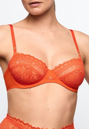 Woman wearing an orange lace bra with thin adjustable straps and a small metal decorative detail at the center front.