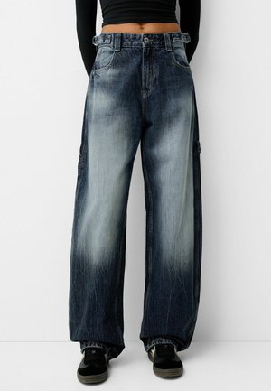 Persona vistiendo jeans azul deslavado de tiro alto y pierna ancha, zapatillas negras, y una camiseta negra de manga larga recortada, de pie frente a un fondo blanco.