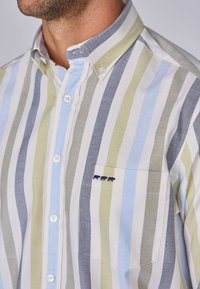 Homme portant une chemise rayée à boutons avec des rayures verticales bleu, vert, gris et blanc, ainsi qu'un petit logo brodé au-dessus de la poche poitrine.
