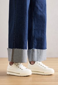 Adidași albi cu o talpă groasă, suprafață texturată și ochiuri aurii, asortați cu pantaloni din denim largi, tăiați, cu margine desfăcută.