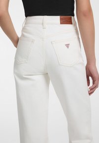 Jeans de mezclilla blancos con cintura alta, bolsillos traseros y una etiqueta de marca marrón. Textura suave y diseño clásico de cinco bolsillos.