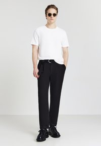 Uomo che indossa occhiali da sole, camicia bianca a maniche corte, pantaloni neri con cintura e scarpe nere, in piedi su uno sfondo bianco uniforme.