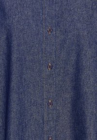 Chemise en denim bleu présentant un tissu texturé, une fermeture boutonnée avec des boutons foncés et des détails de surpiqûres contrastantes le long de la patte de boutonnage.
