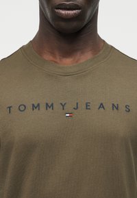 Κοντινό πλάνο ατόμου που φοράει μπλουζάκι Tommy Jeans σε ελιά πράσινο χρώμα με σκούρο μπλε κείμενο και μικρό κόκκινο, λευκό και μπλε λογότυπο στο στήθος.