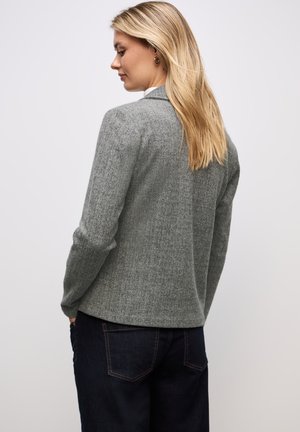Blonde Frau, die ein graues Fischgratblazer und dunkle Jeans trägt, mit den Händen in den Taschen stehend, nach links auf einem schlichten weißen Hintergrund blickend.