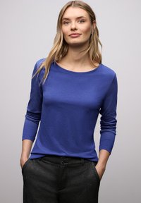 Niebieska koszula z długim rękawem wykonana z gładkiego, lekkiego materiału. Posiada okrągły dekolt i dopasowane mankiety, zestawiona z ciemnymi, fakturowanymi spodniami.