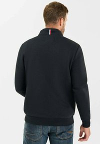 Uomo di spalle che indossa un maglione nero a maniche lunghe con una piccola striscia rossa, bianca e blu sul colletto e jeans blu.