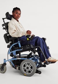 Elektrische rolstoel met een blauw frame, grote grijze wielen en een comfortabele zwarte stoel. De gebruiker draagt een gestreept shirt en een blauwe broek.