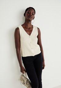 Knit cream sleeveless cardigan with a V-neck, five-button closure, and ribbed texture, paired with black fitted jeans and holding a bag.