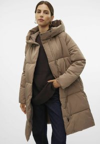 Abrigo acolchado marrón con capucha extragrande, diseño acolchado, cierres a presión y bolsillos laterales, llevado sobre un suéter oscuro y jeans azules.