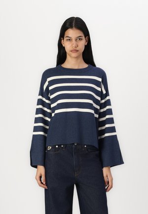 Suéter a rayas azul marino con rayas horizontales blancas, textura acanalada, mangas anchas y un corte relajado, combinado con jeans oscuros.