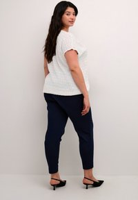 Haut blanc avec un motif bleu clair, manches courtes, associé à un pantalon ajusté bleu marine. Le mannequin porte des escarpins noirs à bride arrière.