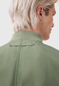 Green bomber jacket featuring a ribbed collar, smooth fabric, and contrasting seam details. Back view highlights the collar and material texture.