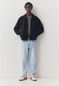 Jonge man die met zijn handen in de zakken van een zwart jack staat, draagt een grijs shirt, lichtblauwe spijkerbroek en witte sneakers, tegen een effen muur.