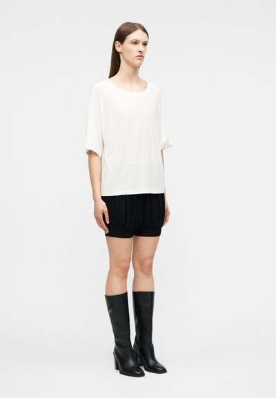 Γυναίκα στέκεται με φαρδιά λευκή μπλούζα με κοντά μανίκια, μαύρα σορτς και μαύρες ψηλές μπότες με τακούνι μέχρι το γόνατο, μπροστά σε απλό φόντο.
