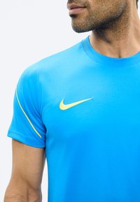 Man draagt een felblauw Nike sportshirt met een gele swoosh-logo, kijkt opzij, tegen een effen lichte achtergrond.
