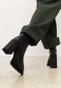 Bottines en cuir noir avec un talon carré et une semelle en caoutchouc texturée, associées à un pantalon vert. Fini lisse et design épuré.