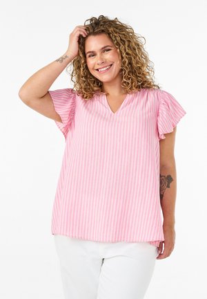 Mujer con cabello rizado que lleva una blusa de manga corta a rayas rosas y blancas y pantalones blancos, sonriendo con una mano tocándose el cabello.