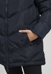 Marineblaue Daunenjacke mit gesteppter Musterung, Reißverschluss und Seitentasche. Hergestellt aus glattem, leichtem Material mit einer sanften Textur.