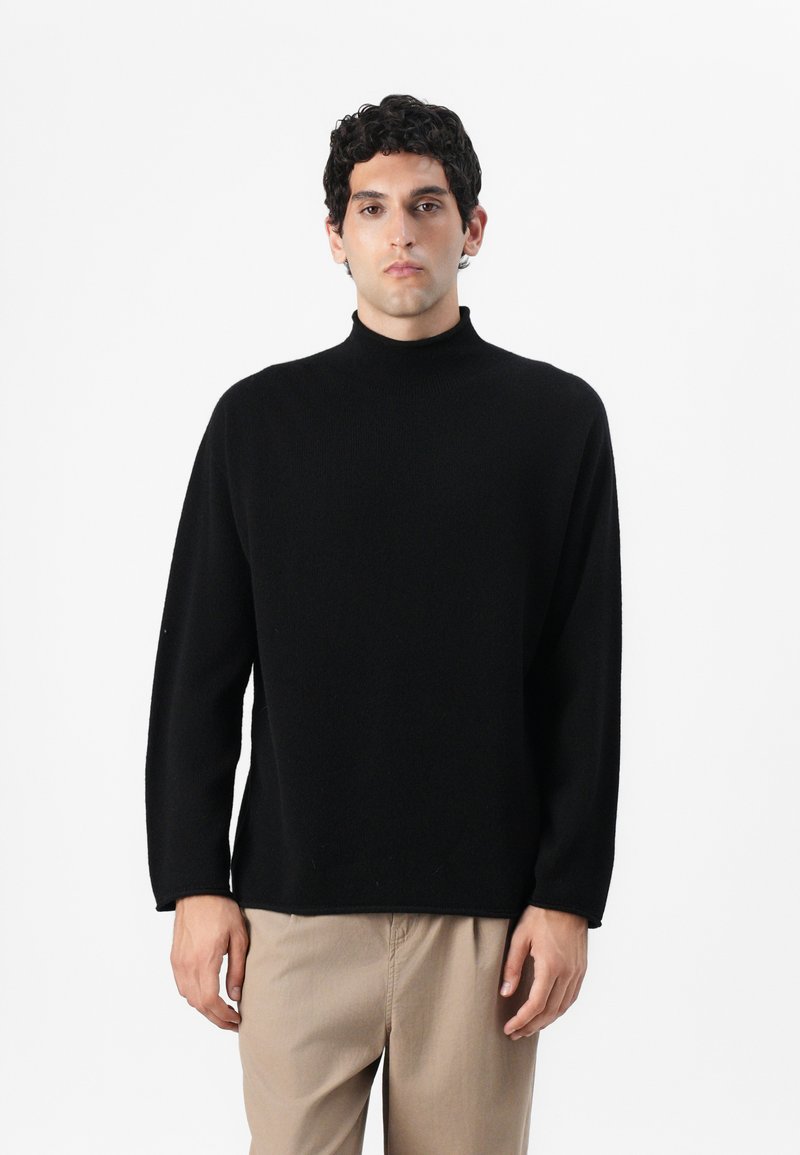 Maglione a collo alto di colore nero con vestibilità comoda, maniche lunghe e texture morbida; abbinato a pantaloni di colore chiaro.