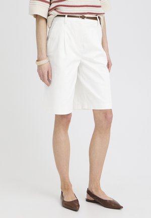 Kvinde iført hvide skræddersyede knælange shorts, brunt bælte, stribet striktop, brune spidse flade sko og beige armbånd, stående mod en hvid baggrund.