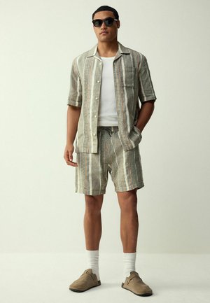 Man wearing sunglasses, a striped short-sleeve button shirt, matching shorts, white socks, and brown slip-on shoes standing against a plain background.