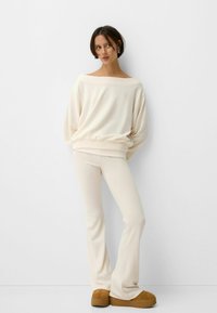 Off-white knit sweatshirt with an off-shoulder neckline, paired with matching flared pants. Soft texture and relaxed fit. Brown platform shoes.