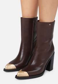 Botas de tornozelo em couro castanho com biqueira metálica dourada, salto largo e bloco, e fecho lateral para fácil calço. Textura suave por toda a parte.