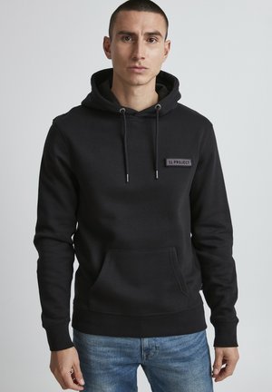 Hoodie noir avec une poche avant et des cordons de serrage. Présente un empiècement texturé portant l'étiquette "11 PROJECT" sur la poitrine. Tissu doux, coupe décontractée.