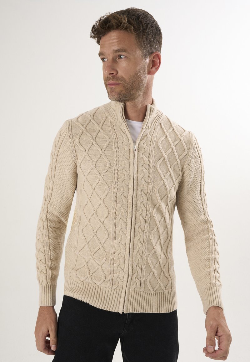 Maglione beige con zip, caratterizzato da un motivo a treccia testurizzato, polsini e orlo a costine, e un design con colletto alto.