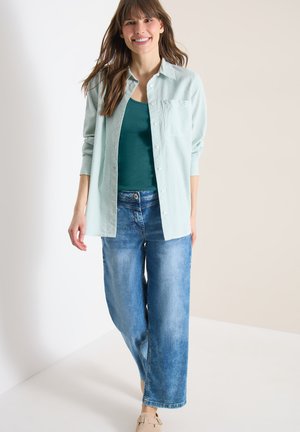 Ung kvinde smilende, iført en lysegrønstribet skjorte over en mørkegrøn top, blå jeans med vide ben og beige sko, stående i et lyst studie.