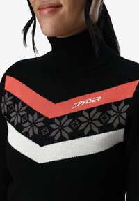 Pull en col roulé noir avec un motif en chevrons rouge, blanc et gris, et des motifs floraux sur la poitrine. Logo "SPYDER" à l'avant.
