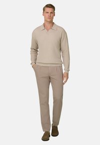 Homme portant un polo en maille beige à manches longues, un pantalon marron clair et des mocassins en daim marron, debout avec une main dans la poche sur fond blanc.