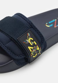 Chancla de color marino con una correa de tela, que presenta un parche de logo colorido y una suela texturizada. La correa cuenta con un cierre de Velcro para mayor ajustabilidad.