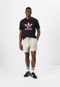 Svart t-skjorte med rosa Adidas-logo, lyse mønstrede shorts, svart sko og stripete sokker, stående mot en enkel hvit bakgrunn.