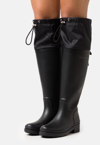 Bottes de pluie noires en caoutchouc avec une semelle texturée, un haut tige, et un haut en nylon noir ajustable tiré par des cordons. Logo sur le côté.