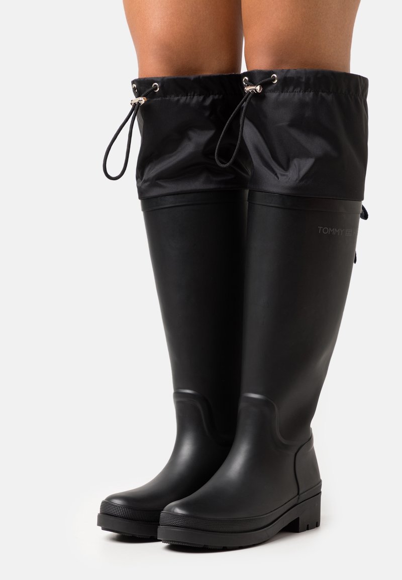 Bottes de pluie noires en caoutchouc avec une semelle texturée, un haut tige, et un haut en nylon noir ajustable tiré par des cordons. Logo sur le côté.